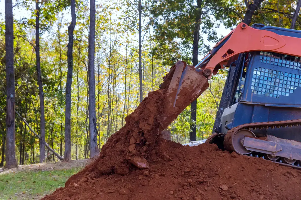 Compact track loader dumping red clay on a wooded lot
